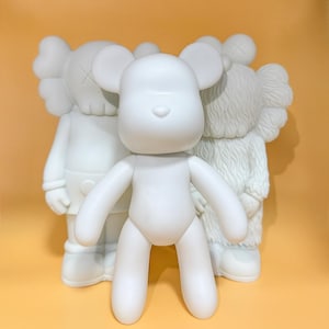 Könnte beinhalten: Drei weiße Vinyl-Spielfiguren. Die zentrale Figur ist ein bärenähnliches Design mit abgerundeten Gliedmaßen und Ohren. Die Figuren dahinter haben ähnliche Merkmale, darunter ein kreuzäugiges Gesicht und eine pelzige Textur. Die Spielzeuge stehen vor einem gelben Hintergrund.