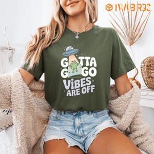 May include: An olive green short-sleeve t-shirt featuring a graphic of a green frog being abducted by a blue and silver UFO. The text reads "GOTTA GO VIBES ARE OFF" in white and light purple lettering, accompanied by small white star accents.