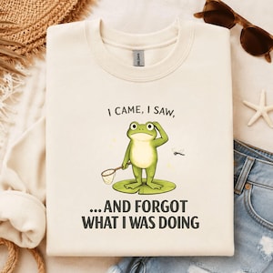 May include: A cream-colored crewneck sweatshirt featuring a graphic of a green frog standing on a lily pad, holding a small net. The text above the frog reads I CAME, I SAW, and the text below reads ...AND FORGOT WHAT I WAS DOING.