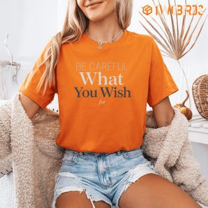 以下が含まれることがあります： 鮮やかなオレンジ色の半袖Tシャツで、白とダークグレーの文字で「BE CAREFUL What You Wish for」とプリントされています。Tシャツはクルーネックで、裾がほつれたライトブルーのデニムショートパンツと組み合わされています。