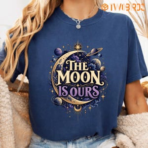 Könnte beinhalten: Ein marineblaues Kurzarm-T-Shirt mit einem Grafikdruck aus einer Mondsichel, Planeten und Sternen. Der Text "THE MOON IS OURS" ist mittig in einer dekorativen Schriftart aufgedruckt. Das Design enthält violette Wolken und goldene Akzente.