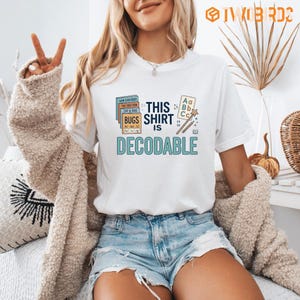 Op de afbeelding: Een wit t-shirt met korte mouwen en een grafische print met de tekst THIS SHIRT IS DECODABLE, vergezeld van illustraties van gestapelde boeken, een kleine alfabetkaart en een potlood, uitgevoerd in blauw-, oranje- en bruintinten.