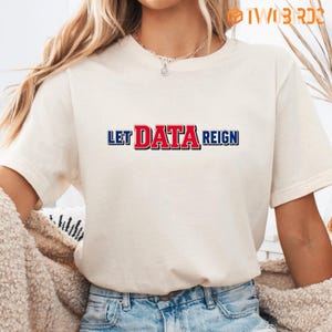 Puede incluir: Una camiseta de manga corta de color crema con el texto LET DATA REIGN impreso en el pecho. La palabra DATA está en letras rojas en negrita con un borde negro, mientras que las palabras LET y REIGN están en letras azules con borde negro.