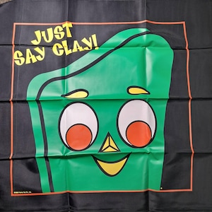 May include: A black square fabric banner featuring a large green character with white eyes, orange pupils, a yellow triangular nose, and a yellow mouth. The text "JUST SAY CLAY!" is printed in yellow in the upper left corner within an orange border.
