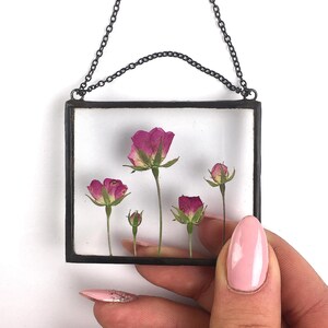 Op de afbeelding: Een ketting met een zwart metalen frame en een glazen insert met geperste roze bloemen. De bloemen zijn in een cluster gerangschikt met stengels die naar boven reiken.