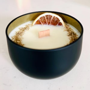 May include: A black candle in a round container with a gold rim. The candle has a white wax with a wooden wick and is decorated with dried citrus and herbs. The candle is a decorative home fragrance item.