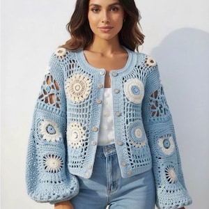 Könnte beinhalten: Hellblauer Häkelcardigan mit cremefarbenen Blumenmustern. Der Cardigan hat eine Knopfleiste, lange Ärmel und einen runden Ausschnitt. Die Ärmel sind ballonförmig. Das Häkeldesign ist offen und luftig, mit einem zarten Muster.