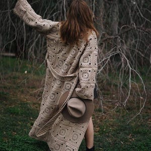 Könnte beinhalten: Ein langer, beigefarbener Häkelmantel mit geometrischem Muster und passendem Gürtel. Ein brauner Hut wird in der Hand gehalten. Die Person geht auf grünem Gras mit kahlen Bäumen im Hintergrund. Der Mantel ist etwa 140 cm lang.