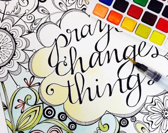May include: A hand-lettered inspirational quote, "pray changes things", in black ink on a white background. The quote is surrounded by a yellow watercolor cloud and a black and white floral design. A watercolor palette and a paintbrush are visible in the top right corner.