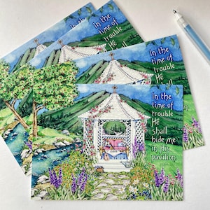 May include: Watercolor art prints featuring a gazebo scene with the text "In the time of trouble He shall hide me in His pavilion." The prints showcase a gazebo, flowers, a stream, and mountains, with a pen in the background.