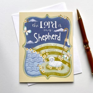 May include: Greeting card with the text "the Lord is my Shepherd" in a decorative frame. The card features a lamb, a shepherd's staff, butterflies, and the phrases "He makes me lie down in green pastures" and "He leads me beside still waters". A pen rests beside the card.