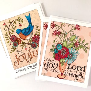 May include: A stack of art prints featuring colorful illustrations. One print shows a bluebird perched on a floral branch with the words "be joyful." Another print displays a floral bouquet in a vase with the text "the joy of the Lord is my strength."