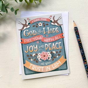 May include: Greeting card with the text "MAY THE GOD of HOPE FILL YOU WITH ALL JOY and PEACE as you TRUST IN HIM". The card has a teal background with floral and bird illustrations. A patterned pencil rests on the side.