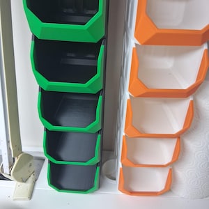 May include: Vertical planters in green and orange. The green planter has four stacked, open-top compartments with black interiors. The orange planter has three stacked compartments with white interiors. Both planters are made of plastic and are designed for vertical gardening.