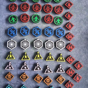 May include: A collection of game tokens in various shapes and colours. The set includes circular green and red tokens with figure icons, blue hexagonal tokens with droplet icons, white hexagonal tokens with web patterns, and triangular yellow and pink tokens.