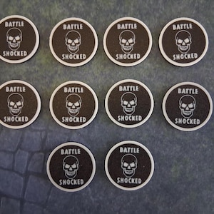 May include: Ten circular tokens with a black textured surface and a white border. Each token features a white graphic of a skull in the centre with the word BATTLE printed above and the word SHOCKED printed below the skull in white text.