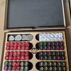 May include: A wooden box with compartments for storing dice. The box has a black foam lining and a Scoffcorp logo. The dice are in various colours, including red, green, and black. There are also silver tokens in the box.