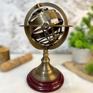 Puede incluir: Una esfera armilar de latón con signos del zodiaco, incluyendo Piscis y Aries, montada sobre una base de madera rojo oscuro. La esfera tiene un diseño antiguo con marcas de medición.