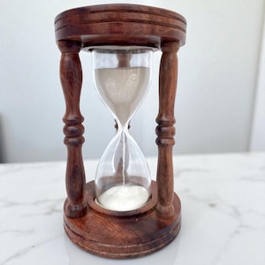 May include: A wooden hourglass with a dark brown finish. The hourglass has a clear glass center filled with light-colored sand. The wooden frame features decorative turned columns and a circular base and top.
