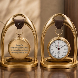 May include: Two gold-toned metal desk clocks suspended from arched stands by brown leather straps. One clock face features Roman numerals and the text "Frankland Murphy EST 1849 OXFORD". The other side displays an engraved award inscription.