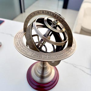 Puede incluir: Una esfera armilar de color bronce con una base roja oscura. La esfera tiene múltiples anillos con números y texto, incluyendo "Tripoly" y "Zaba Isle". El diseño es intrincado y detallado, lo que sugiere un tema celestial o científico.