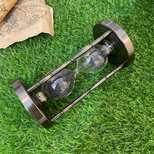 May include: An antique-style hourglass with a dark metal frame and base. The glass bulbs contain black glitter-like sand. The hourglass is designed with a vintage aesthetic, suitable for home decor or as a unique gift.