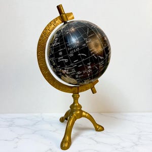 Puede incluir: Un globo terráqueo negro decorativo con detalles metálicos dorados. El globo presenta marcas geográficas detalladas y está montado sobre un soporte de tres patas. El soporte tiene un meridiano circular y una barra horizontal en la parte superior.
