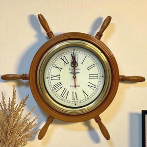 Puede incluir: Un reloj de pared redondo con forma de timón de barco de madera y marco color latón. La esfera de color blanco roto presenta números romanos negros, una escala de minutos y el texto "Dos Santos Estd 1747" y "London" con manecillas negras y rojas.
