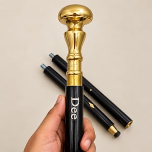 Pode incluir: Uma bengala com um cabo de metal dourado polido com topo arredondado e haste de madeira preta. A haste está gravada com o nome Dee em letras brancas. Dois segmentos adicionais de haste de madeira preta com conectores metálicos são visíveis.