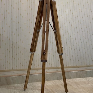 A natural wood tripod easel stand with splayed legs for stability For a plant/decorative stand: "Rustic wooden tripod plant stand with angled legs joined at the top.Vintage-style adjustable tripod stand handcrafted from premium teak wood with polished aluminium and nickel hardware. Features three sturdy wooden legs, an adjustable center column, and a height range of 25 to 50 inches. Ideal for DIY photo booths, DSLR cameras, floor lamps, studio décor, and nautical-inspired interiors.
