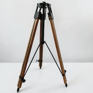 以下が含まれることがあります： 自然な茶色の仕上げの木製脚3本と黒い金属製金具を備えた三脚スタンド。構造には、中央の黒い金属製支柱、調整可能なサポートストラット、および安定性を高めるために各脚の底に尖った足が含まれています。