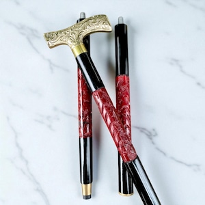 Puede incluir: Bastón decorativo con un mango dorado con un diseño ornamentado. El eje es negro con una sección central roja texturizada. El bastón tiene una punta color latón. Longitud total aproximada de 90 cm.