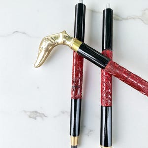 Puede incluir: Tres bastones decorativos. Los bastones tienen una combinación de colores negro y rojo. Los mangos son de color dorado y tienen forma de cabeza de perro. Las secciones rojas están talladas con un patrón floral. Los bastones están sobre una superficie blanca.