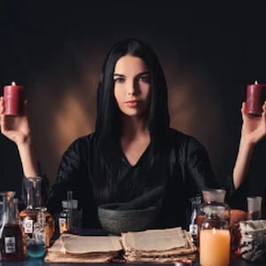 May include: A woman in a black robe holds two red candles in front of her. She is sitting at a table with various bottles, candles, and books. The scene is dark and mysterious, suggesting a ritual or spell casting.