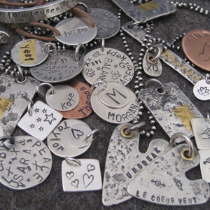 May include: A collection of silver and copper charm pendants with various words and designs, including "Protect Our Earth Mother", "Hope", "Yes", "Peace", "Baby", "Sara", "Taylor", "Thomas", "Kate", "M Morgan", "Unbreak", "Le Coeur Veut", "Kiss My Putt", "Vicki", and "143".