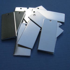 May include: A pile of silver and black metal rectangular tags with holes punched in the top. The tags are arranged in a random pattern.