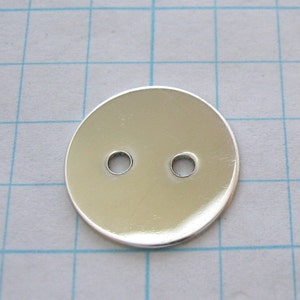 May include: A silver-colored, round button with two holes. The button is smooth and shiny.