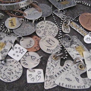 Puede incluir: Una colecci&oacute;n de colgantes de plata y cobre con diversas inscripciones, incluyendo "Protege a nuestra Madre Tierra", "Esperanza", "S&iacute;", "Paz", "Beb&eacute;", "Sara", "Taylor", "Kate", "Thomas", "Morgan", "Inquebrantable", "Le Coeur Veut", "B&eacute;same el culo", "Vicki" y "143".