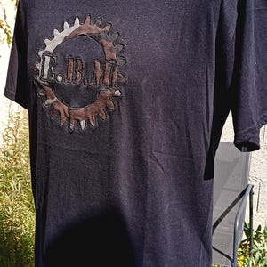 Può includere: T-shirt nero a maniche corte con scollo tondo. La maglietta presenta un design a ingranaggio marrone e grigio con le lettere "E.B.M." al centro. La maglietta è esposta su un manichino.