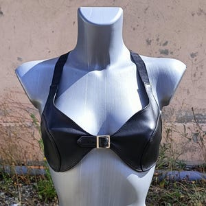 May include: A black leather halter top with a gold-colored buckle closure. The top features a halter neck design with straps that extend over the shoulders. The garment is displayed on a mannequin.