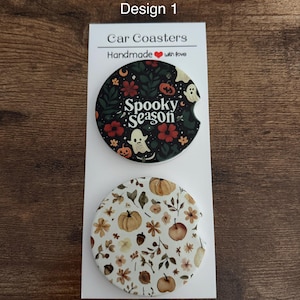 Könnte beinhalten: Zwei runde Auto-Untersetzer auf einer weißen Präsentationskarte mit der Aufschrift Design 1 und Car Coasters Handmade with love. Der obere Untersetzer ist schwarz mit Geistern, Kürbissen und Blumen und trägt den Text Spooky Season. Der untere Untersetzer hat einen weißen Hintergrund mit einem Muster aus Kürbissen, Eicheln und Herbstblättern.