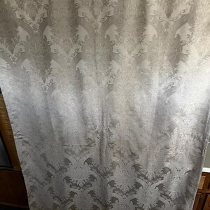 May include: A light-colored curtain with a repeating floral and scroll pattern. The fabric has a subtle sheen, and the design is visible in varying shades of gray and white. The curtain is hanging, with folds visible.