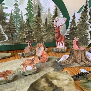 K&ouml;nnte beinhalten: Ein Diorama einer Waldszene mit Papierschnitten von B&auml;umen, Hirschen, F&uuml;chsen, Eulen, Dachsen und Wildschweinen. Der Hintergrund zeigt eine gemalte Waldlandschaft. Die Szene ist auf einer Holzoberfl&auml;che aufgebaut.