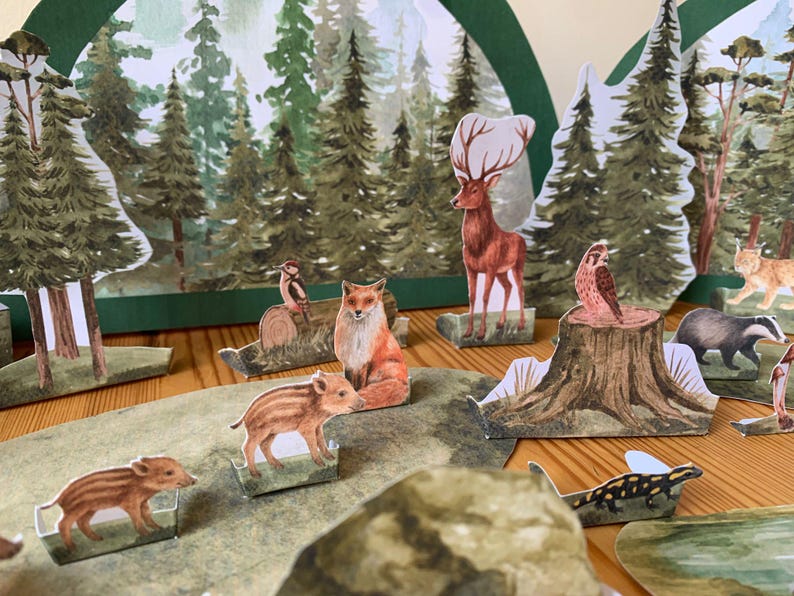 K&ouml;nnte beinhalten: Ein Diorama einer Waldszene mit Papierschnitten von B&auml;umen, Hirschen, F&uuml;chsen, Eulen, Dachsen und Wildschweinen. Der Hintergrund zeigt eine gemalte Waldlandschaft. Die Szene ist auf einer Holzoberfl&auml;che aufgebaut.