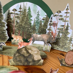K&ouml;nnte beinhalten: Ein Diorama einer Waldszene mit Papierschnitten von B&auml;umen, Tieren und einem Hintergrund aus einem Aquarellwald. Zu den Tieren geh&ouml;ren ein Elch, ein Hirsch, ein Luchs und ein Dachs. Die Szene befindet sich auf einer Holzoberfl&auml;che.