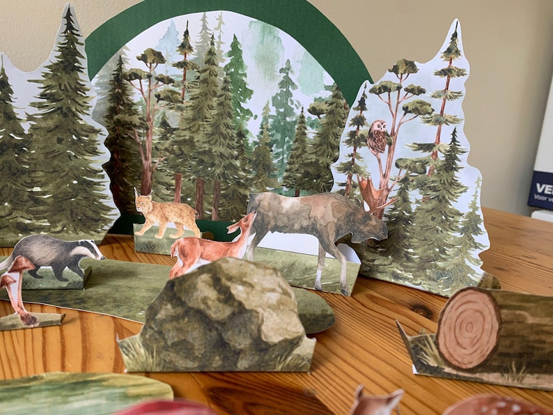 K&ouml;nnte beinhalten: Ein Diorama einer Waldszene mit Papierschnitten von B&auml;umen, Tieren und einem Hintergrund aus einem Aquarellwald. Zu den Tieren geh&ouml;ren ein Elch, ein Hirsch, ein Luchs und ein Dachs. Die Szene befindet sich auf einer Holzoberfl&auml;che.