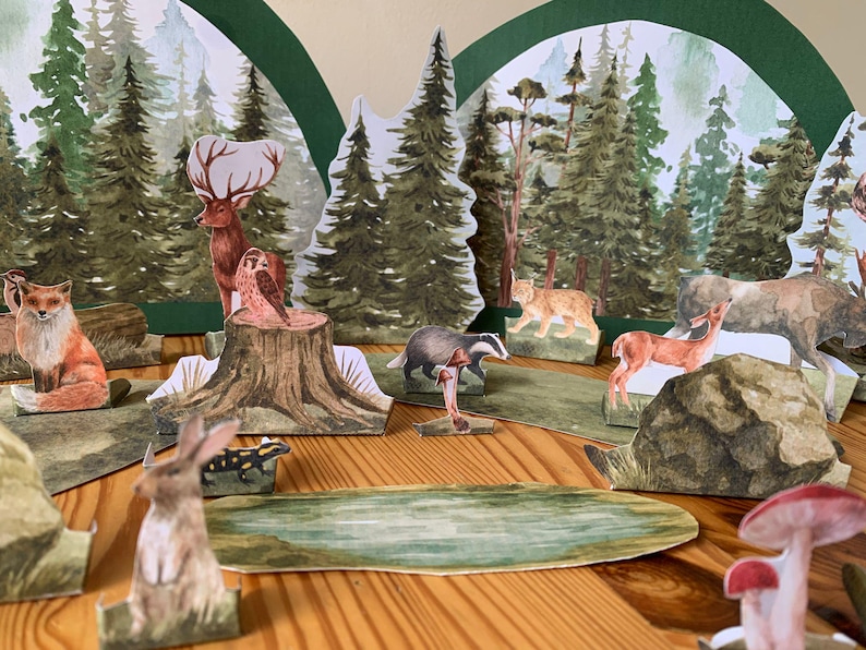 K&ouml;nnte beinhalten: Ein Diorama einer Waldszene mit Papierschnitten verschiedener Tiere, darunter ein Fuchs, ein Hirsch und ein Dachs. Der Hintergrund zeigt gemalte B&auml;ume und einen Teich, wodurch ein dreidimensionaler Effekt entsteht. Die Szene befindet sich auf einer Holzoberfl&auml;che.