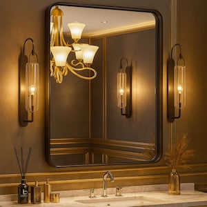 May include: A rectangular black-framed mirror hangs above a marble countertop in a bathroom. Two sconces with clear glass tubes and a gold chandelier provide lighting. A reed diffuser and a vase with dried plants are on the counter.