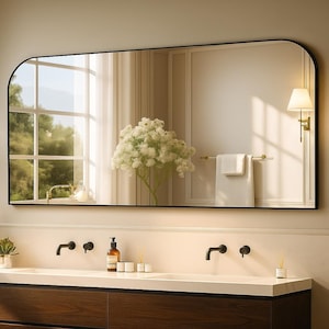 May include: A large, rectangular black-framed mirror with rounded top corners hangs above a double sink vanity. The mirror reflects a window, a floral arrangement, and a wall-mounted light fixture. The vanity has a white countertop and dark wood cabinets.