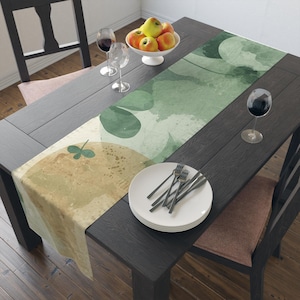 May include: A dark wooden dining table set with a decorative table runner, white plates, and silverware. The table runner features a watercolor design in shades of green and tan. A bowl of apples and wine glasses are also on the table.